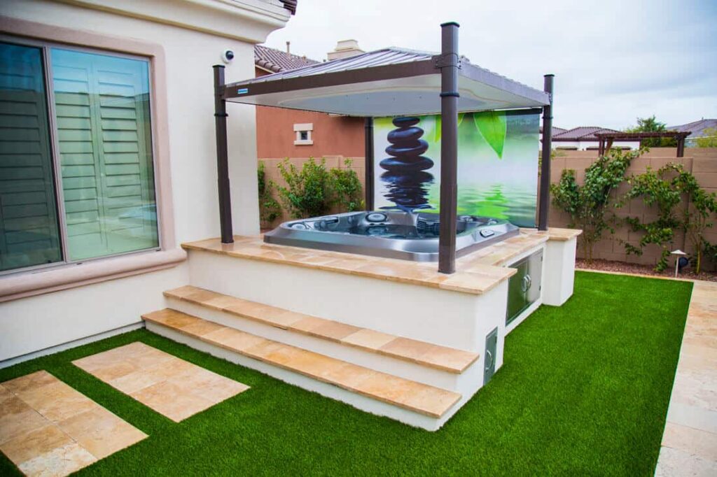 Outdoor patio with a raised hot tub under a four-post pergola, beige tile steps, and artificial grass lawn.
