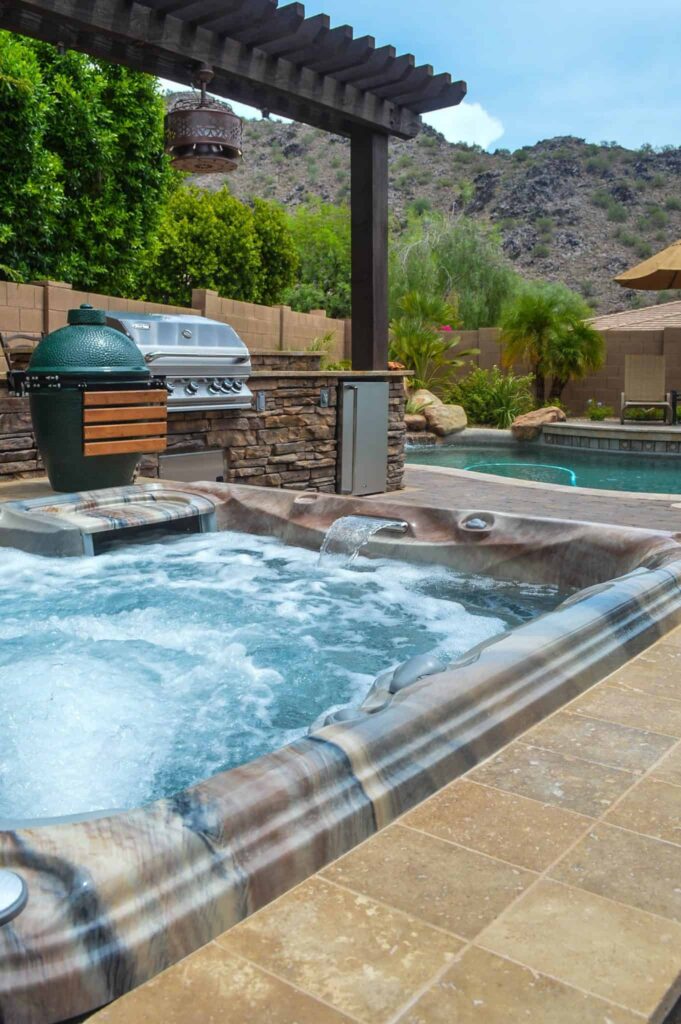 Poolside backyard with a spa, built-in grill, and stone counter under a wooden pergola; desert hills in the background.