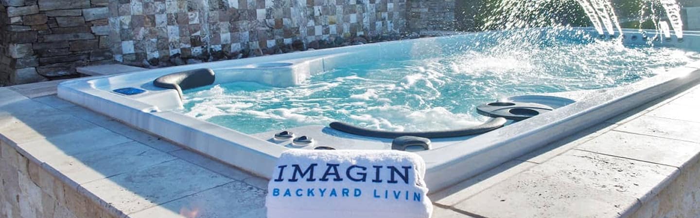 Raised outdoor hot tub on a stone patio with water jets in action and a white towel that reads 'IMAGIN BACKYARD LIVING' draped over the edge
