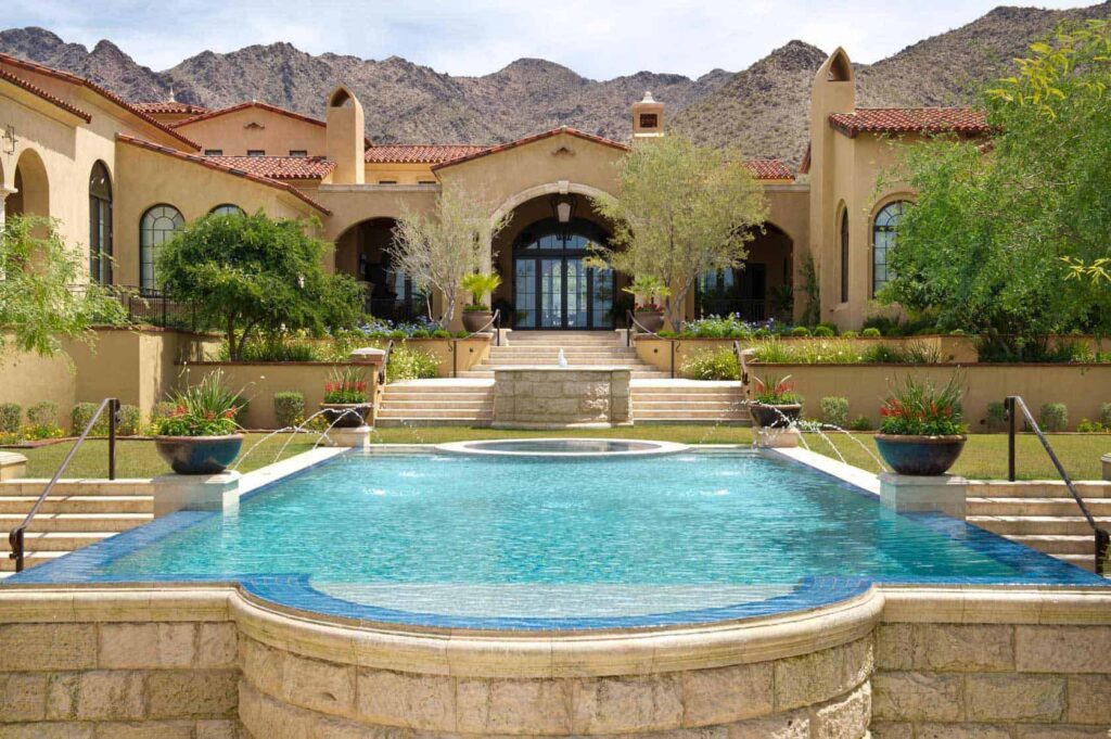 Luxurious villa with a rectangular blue pool and stone fountain, set against desert mountains and landscaped gardens.