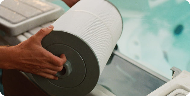 Person handling a large cylindrical pool or spa filter cartridge near a swimming pool while performing maintenance.