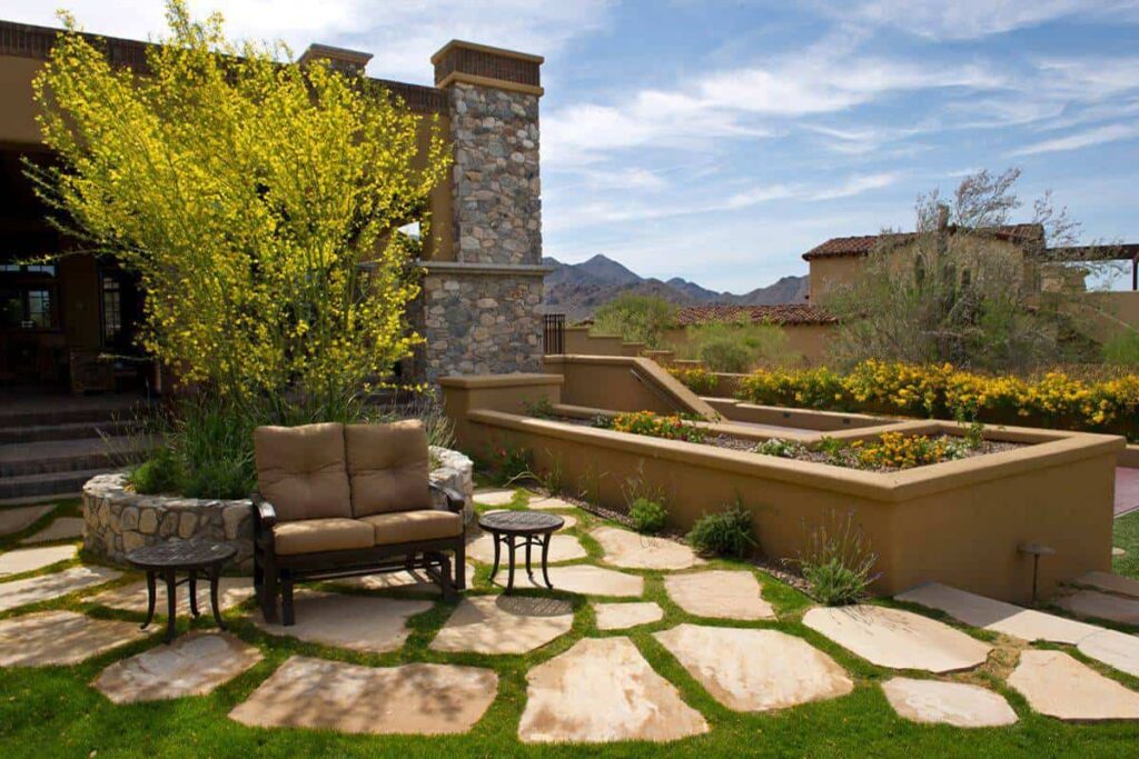 Outdoor seating area with cushioned chairs and small tables beside a stone planter and yellow-flowering tree, in a sunny landscaped yard with mountain backdrop