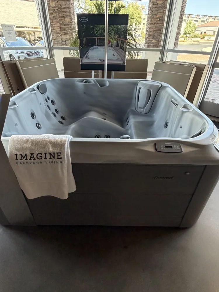 Gray hot tub with multiple jets in a showroom; white towel draped over edge reads IMAGINE Backyard Living.