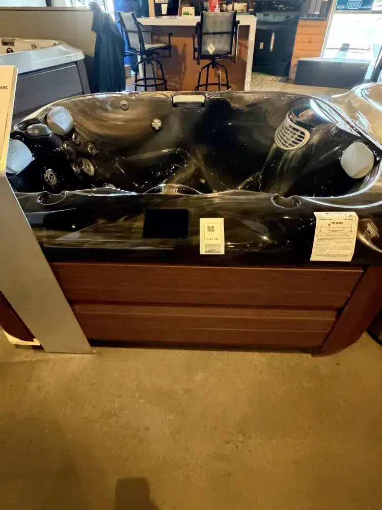 Indoor showroom hot tub with black acrylic interior and wooden base, near a counter and stools; protective plastic film on surface remains intact.