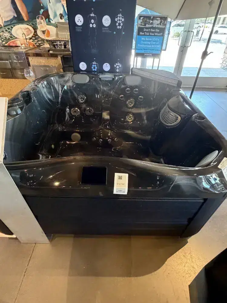 Black hot tub on display with multiple jets and built-in headrests in a showroom near glass doors.