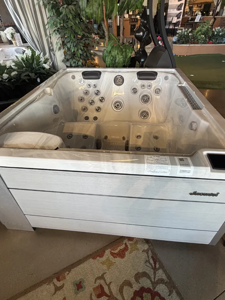 White hot tub with multiple jet nozzles and two built-in headrests on a showroom floor, surrounded by plants and retail displays.