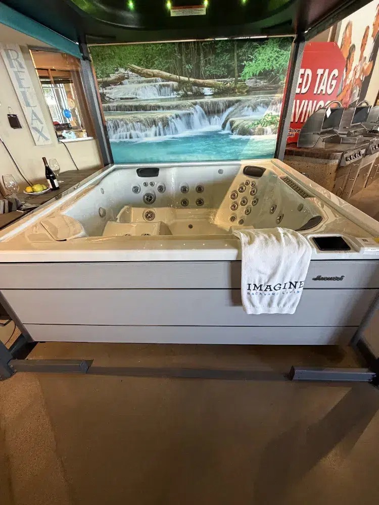 Large indoor showroom hot tub with built‑in jets and a towel draped over the side, backdrop of a waterfall mural behind it.