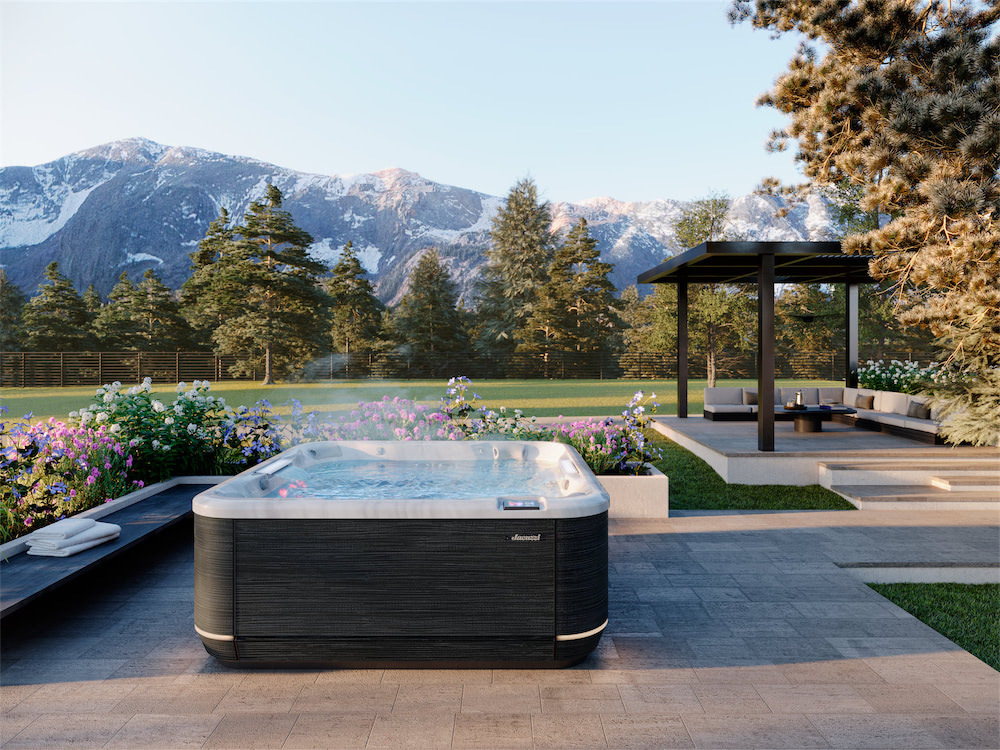 Luxurious backyard scene with a rectangular hot tub on a stone patio, blooming flower beds, and a covered seating area against a backdrop of pine trees and snow-capped mountains.