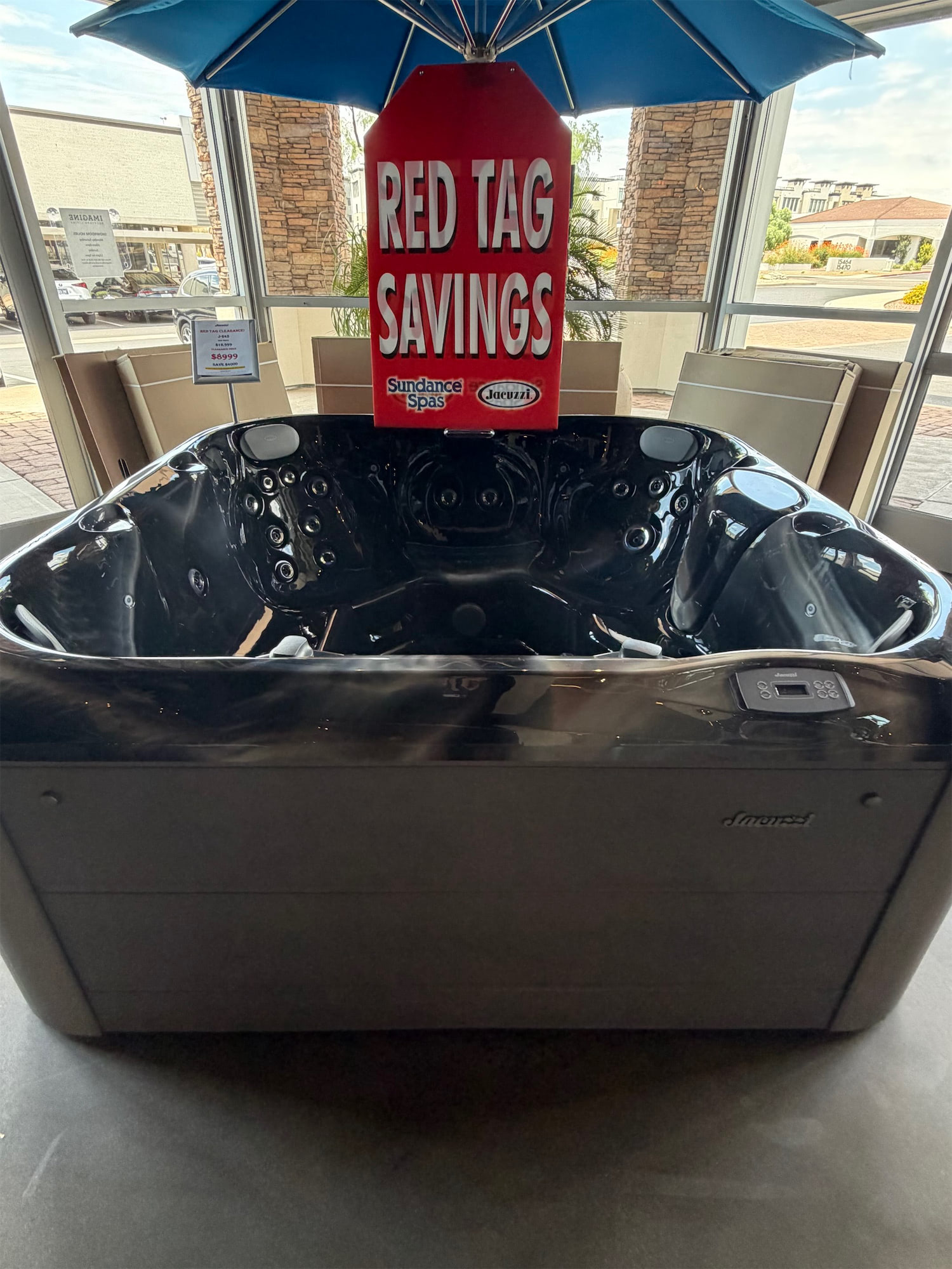 Red promotional sign reading 'RED TAG SAVINGS' displayed over a black hot tub in a showroom, with a blue umbrella overhead and logos for Sundance Spas and Jacuzzi on the sign.