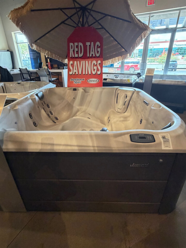 White Jacuzzi hot tub on display in a showroom, under a thatched canopy with a red 'RED TAG SAVINGS' sign overhead.