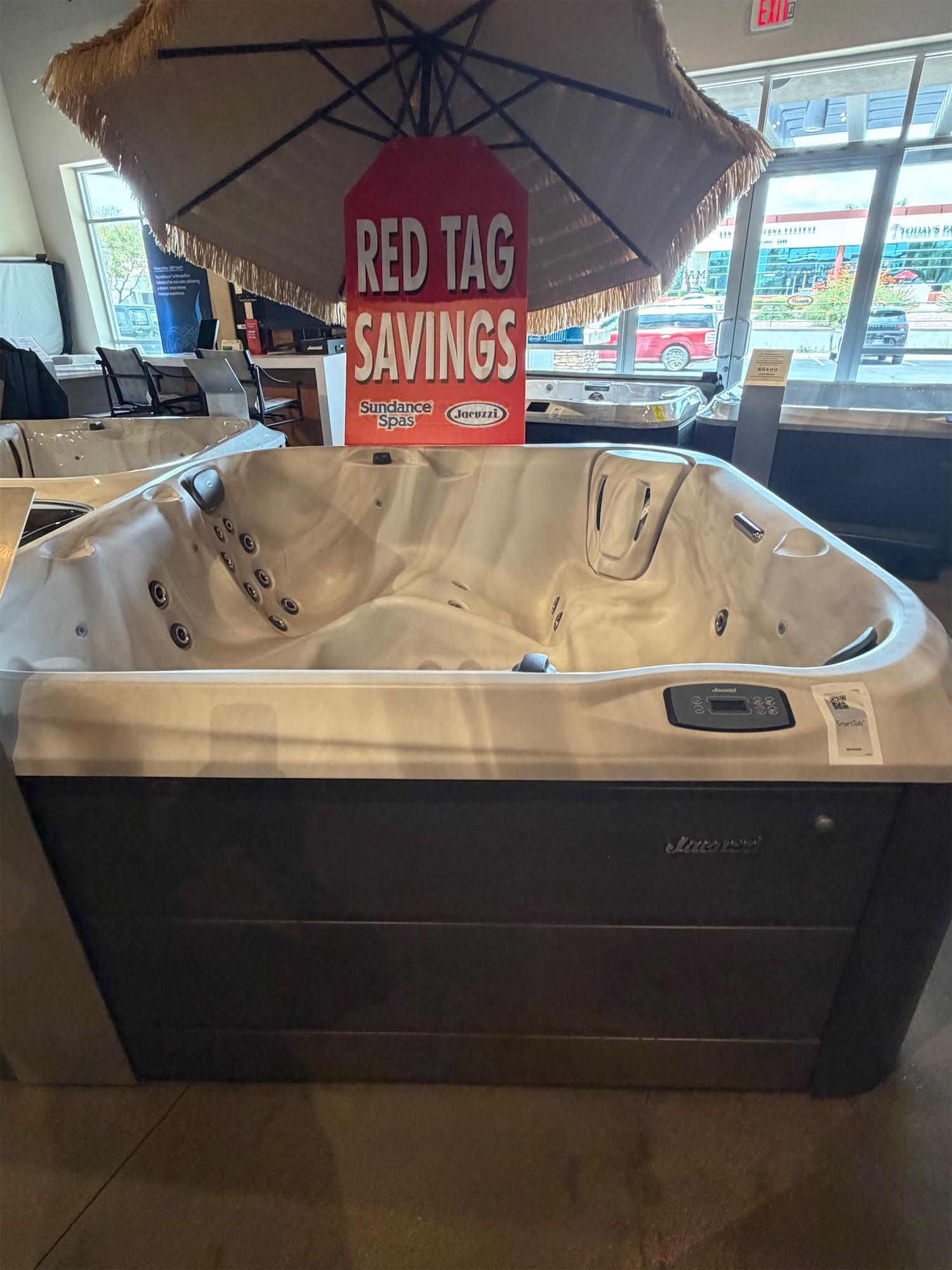 Indoor showroom featuring a large white hot tub with jets, topped by a thatched umbrella and a red 'RED TAG SAVINGS' sign (Sundance Spas/Jacuzzi logos) in the background.