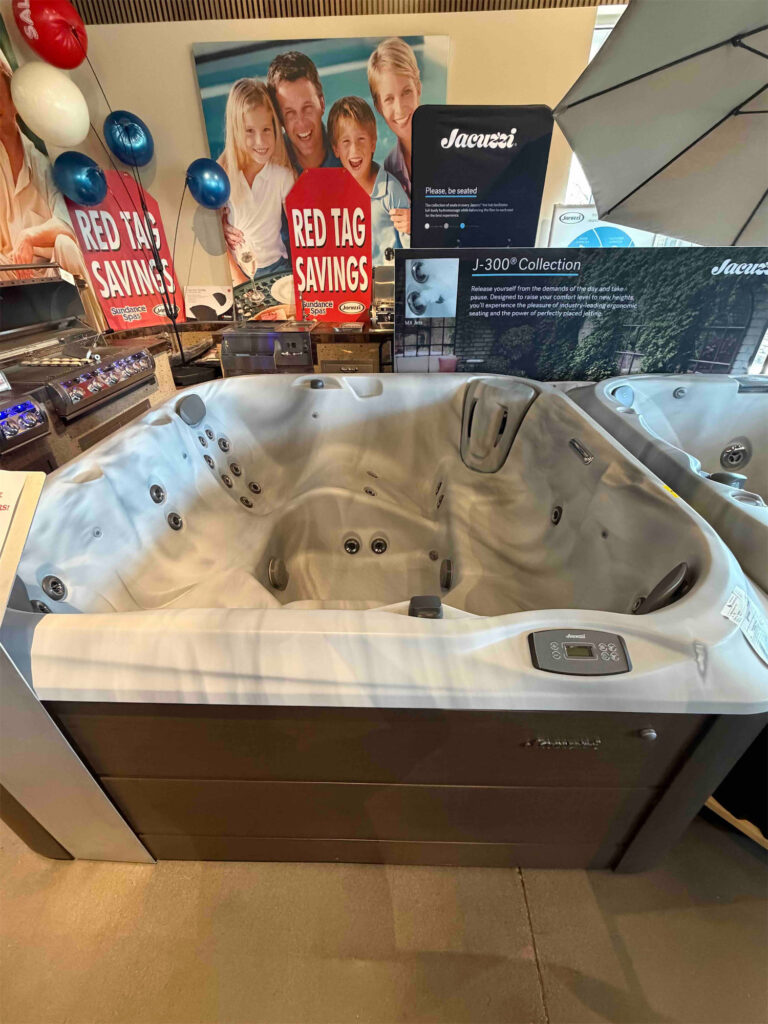 Indoor showroom with a white Jacuzzi hot tub on display, red tag savings posters, and blue balloons in the background.