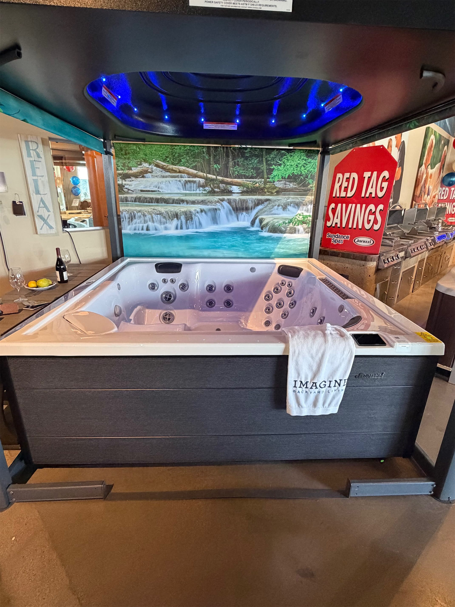 Indoor showroom hot tub with blue LED lights and a nature waterfall backdrop; a 'RED TAG SAVINGS' sign is visible on the right.